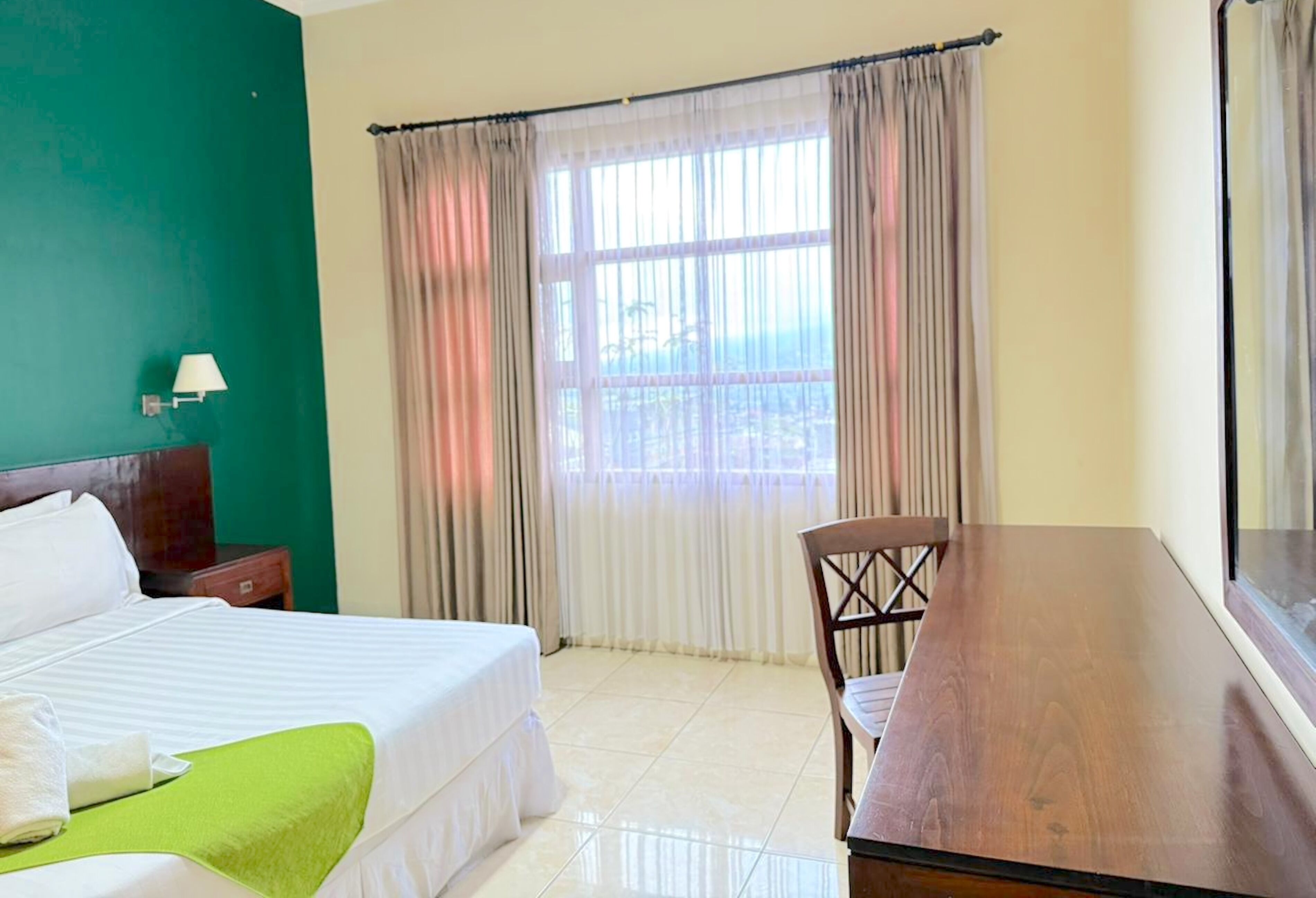 Habitación de hotel con cama, una mesa de madera, una silla, una mesita de noche y ventana con cortinas.