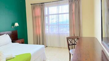 Habitación de hotel con cama, una mesa de madera, una silla, una mesita de noche y ventana con cortinas.