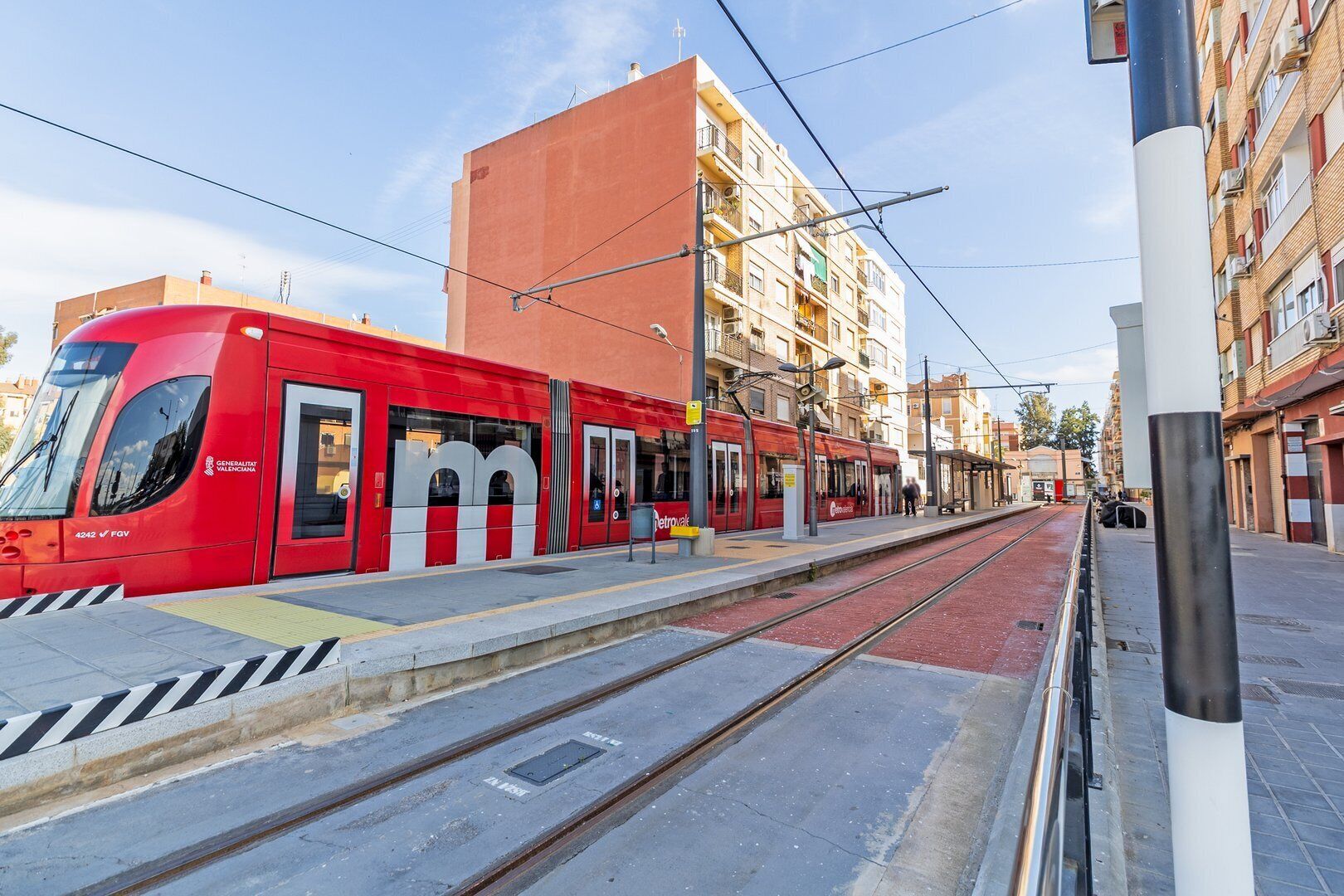 Un tramvia vermell a l'estació, amb cel blau, edificis i una passera per a vianants.