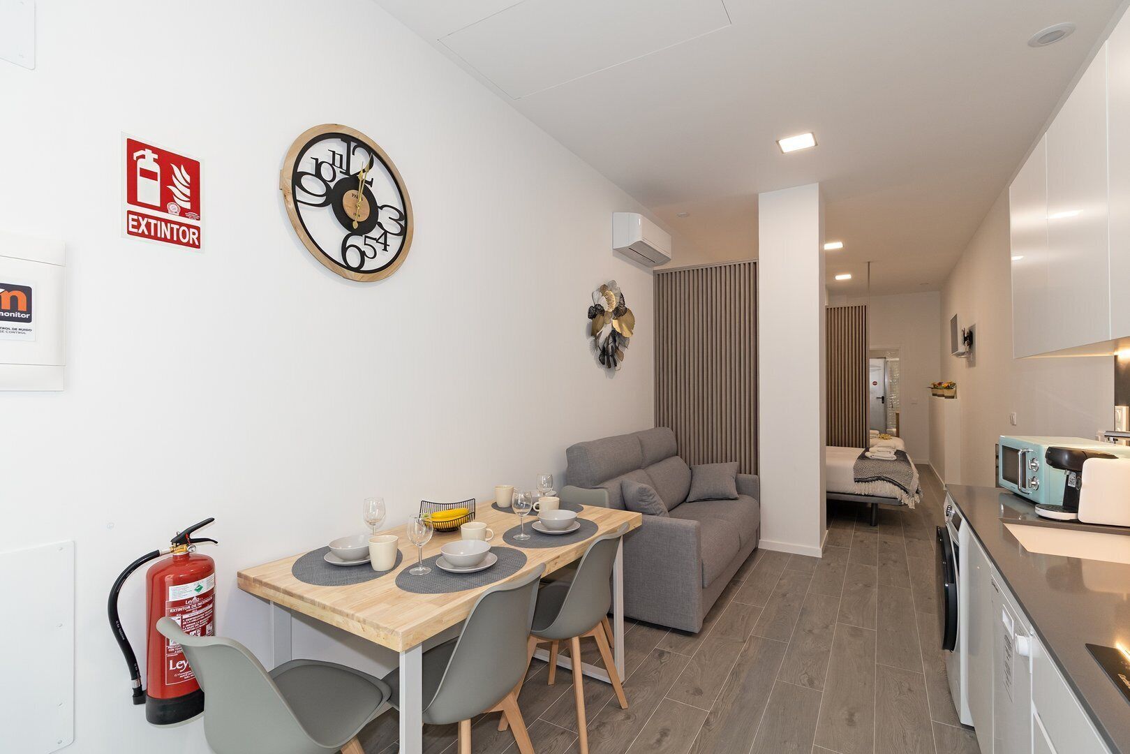 Une chambre d’hôtel moderne avec un coin repas, un canapé gris, une petite cuisine et un extincteur fixé au mur.