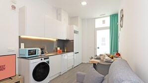 Une cuisine compacte équipée d’un lave-linge, d’un four à micro-ondes et d’une machine à café. On y trouve également un coin repas avec une table pour deux personnes et un canapé gris.