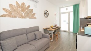 Een moderne woonkamer met een grijze bank, een eethoek met een houten tafel en stoelen, een keuken met een blauw aanrechtblad, en een groot raam met groene gordijnen.