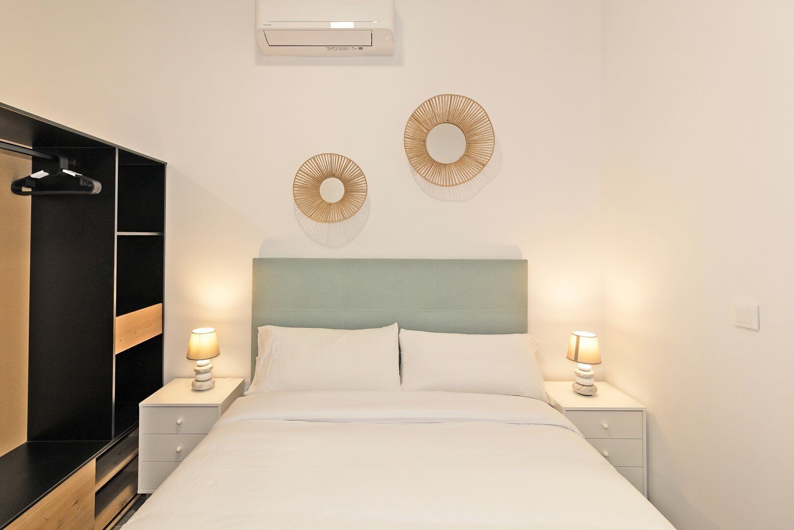 Un dormitorio ordenado con cama, mesitas de noche con lámparas, un armario y dos elementos decorativos en la pared.