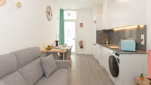 Een moderne keuken met een inbouwoven en een wasmachine, een grijze bank en een eethoek met een tafel voor twee personen.
