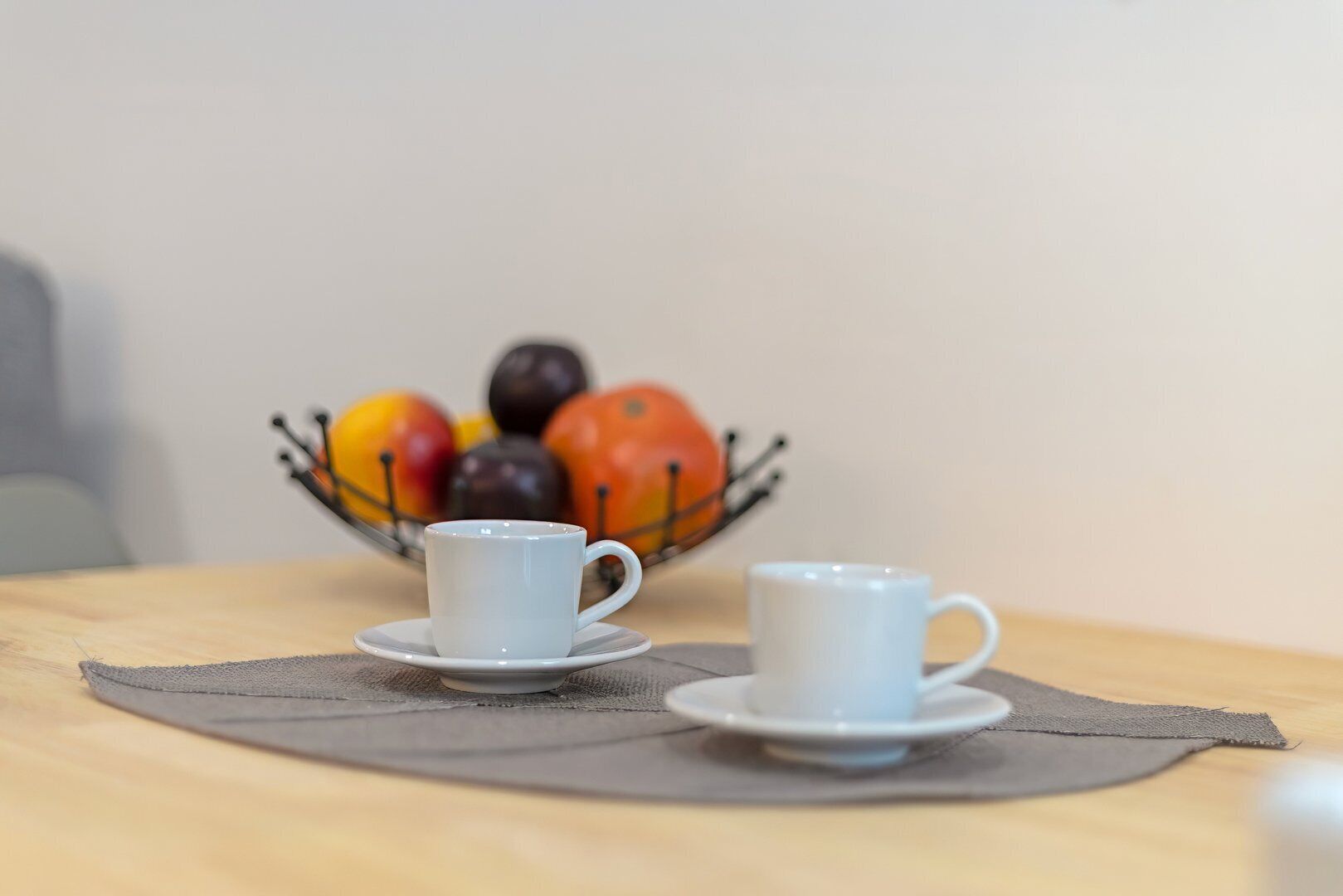 Two white cups on saucers on a table with a gray placemat, and a bowl of fruit in the background.