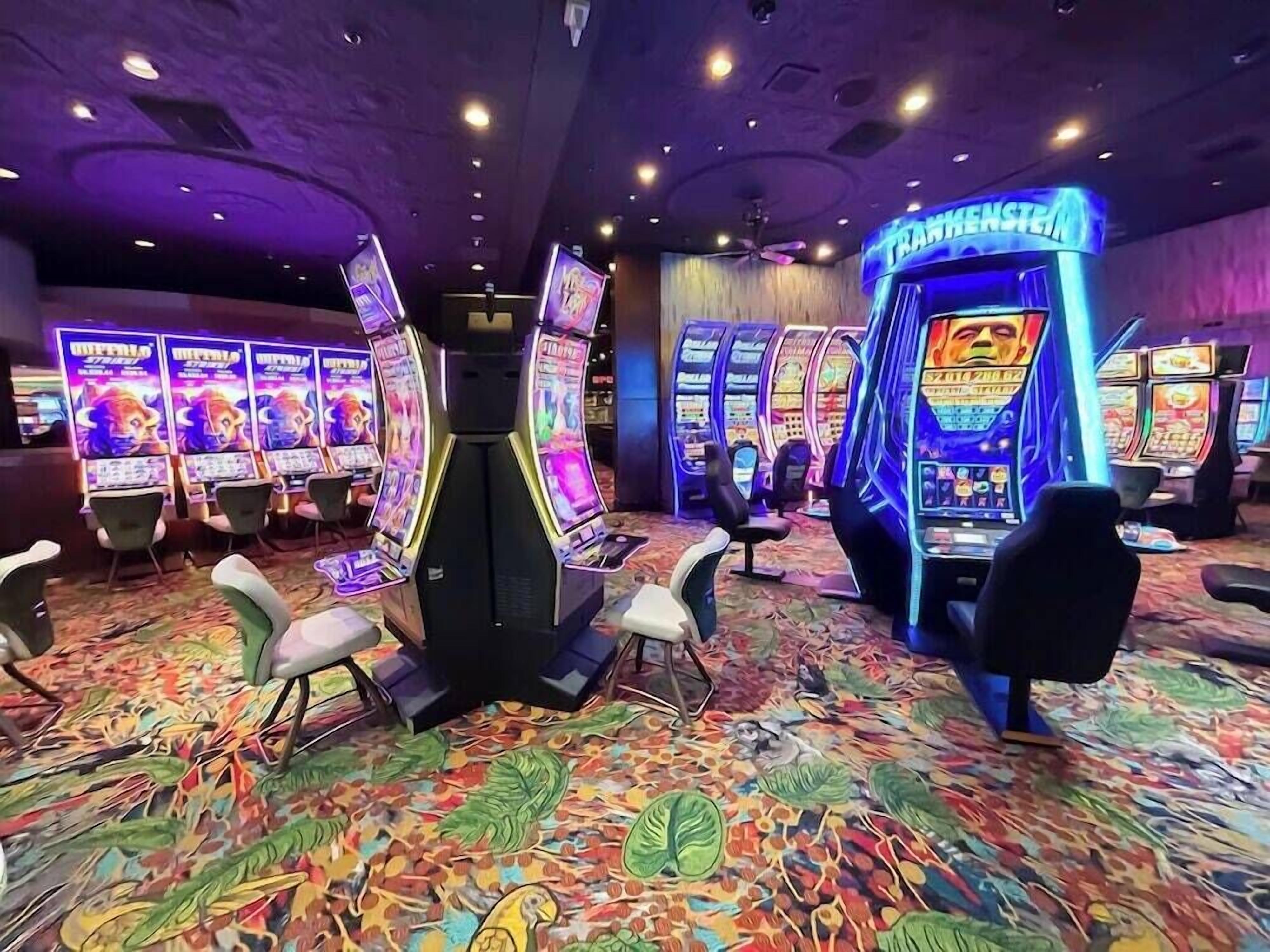 Ett kasino med spelautomater och spelbord, heltäckningsmatta på golvet och belysning i taket.