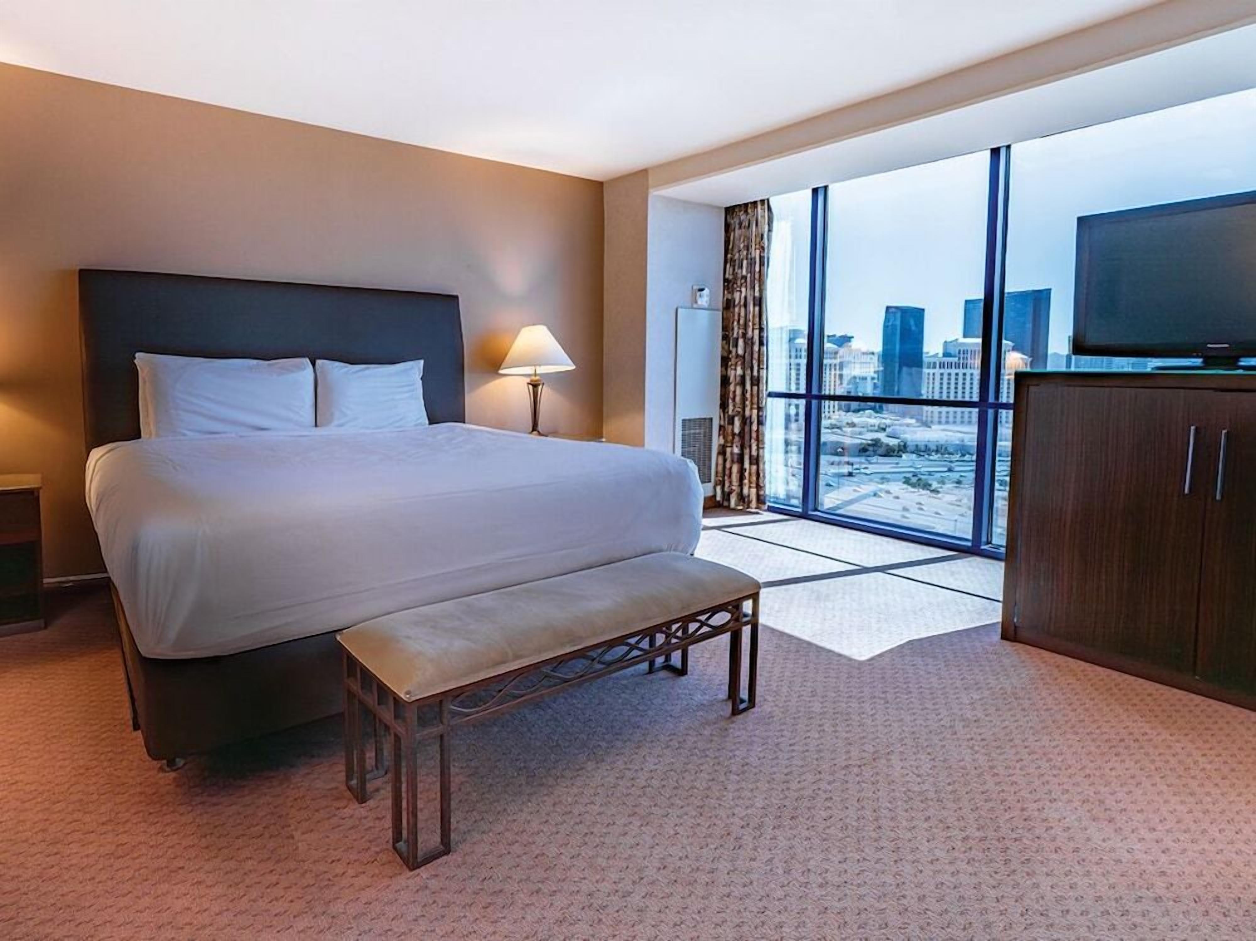 Een hotelkamer met een groot bed, een bankje, een televisie en uitzicht op de stad door grote ramen.
