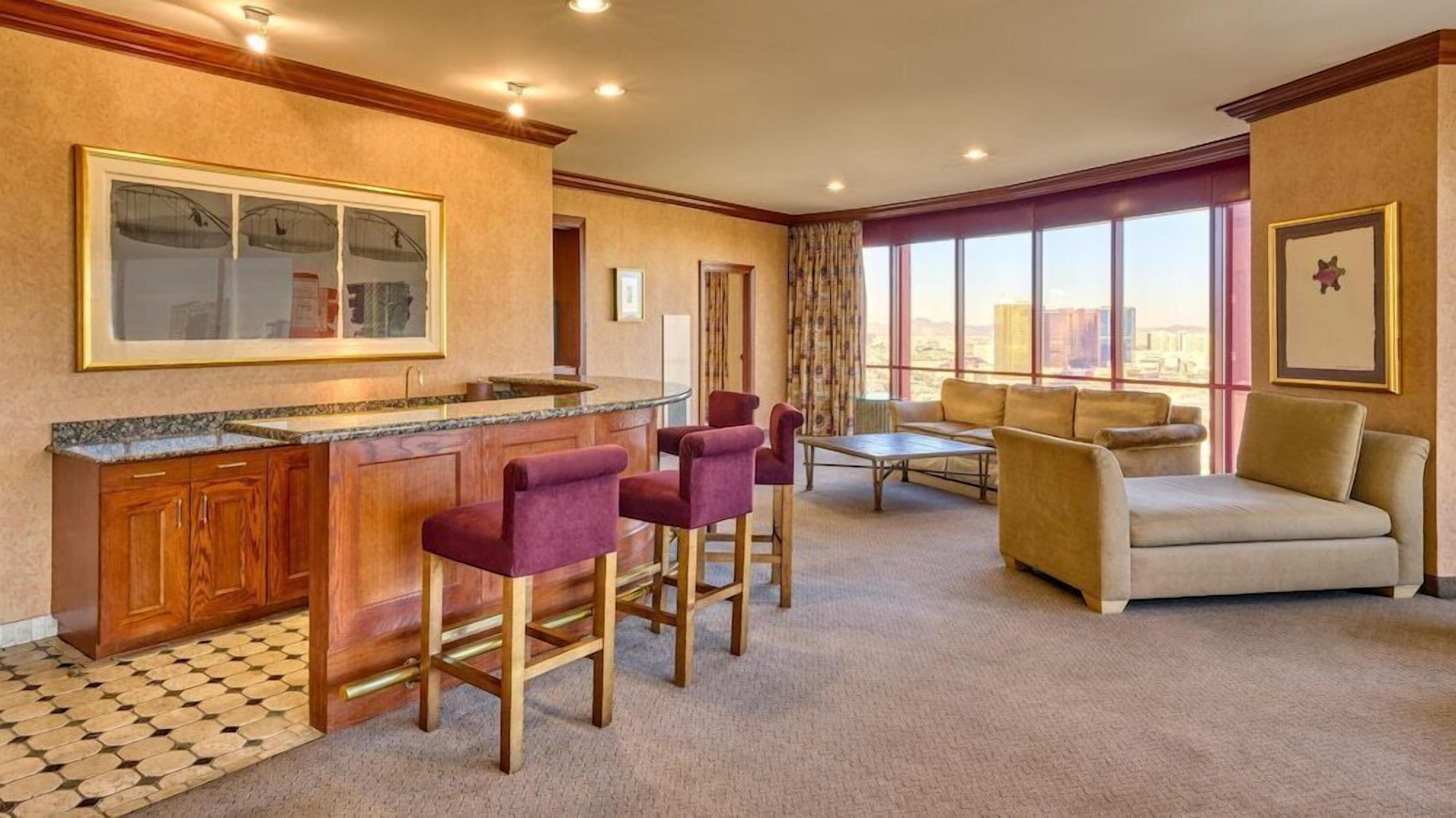 A hotel room with a bar area, a sofa, and a large window offering a city view.