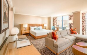 A hotel room with a bed, a sofa, a desk with a lamp, and a view of the city.