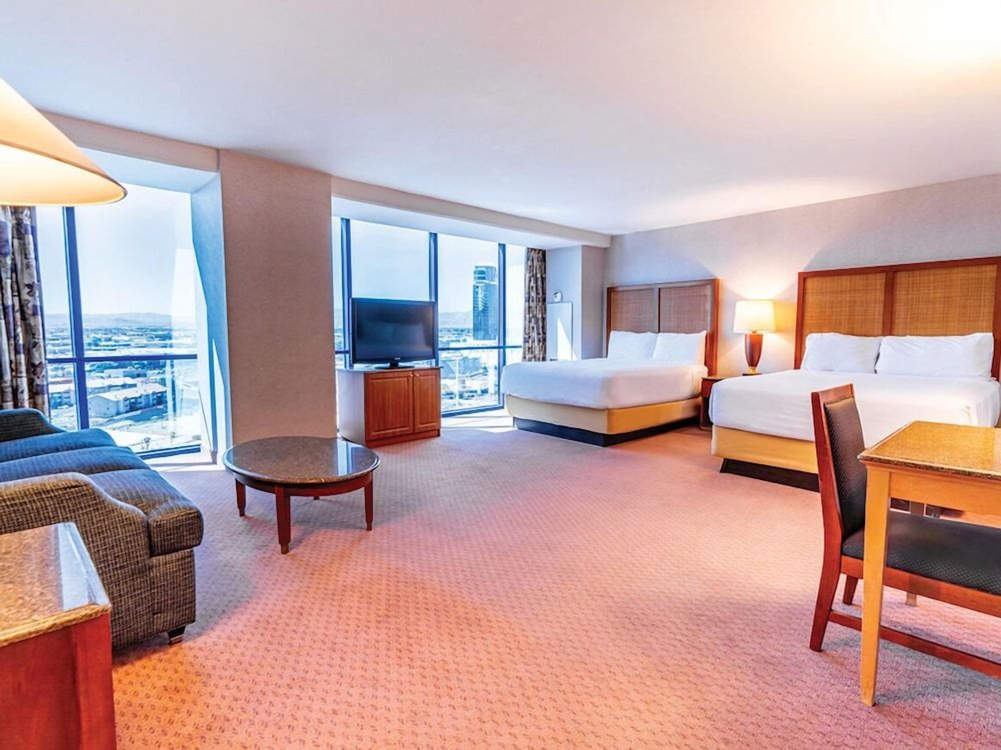 Een hotelkamer met een bed, een bank, een eettafel, een televisie en een groot raam met uitzicht over de stad.