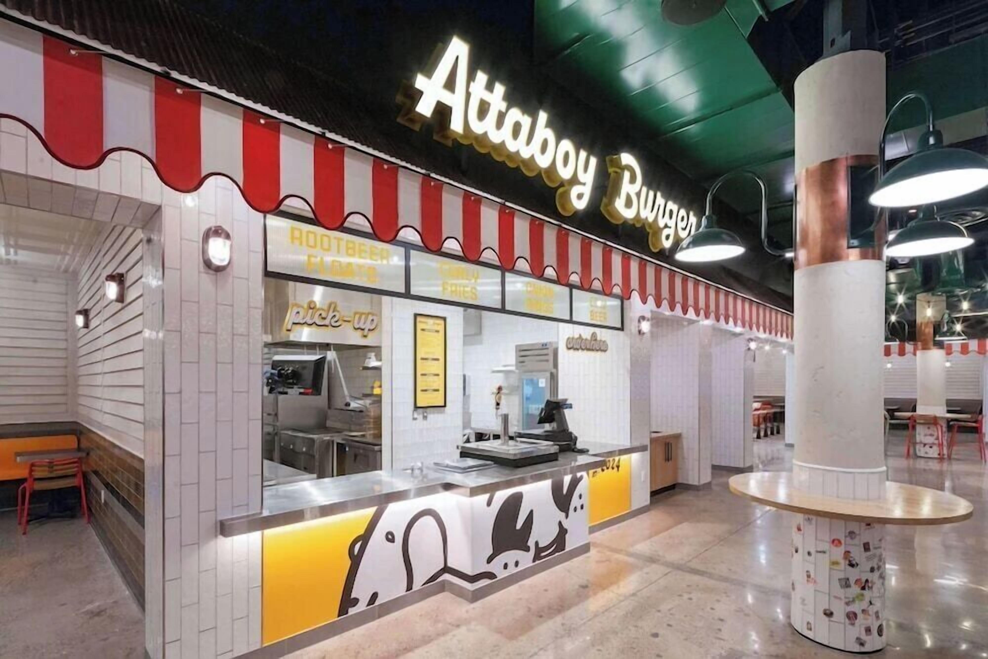 Un patio de comidas con un mostrador iluminado de Attaboy Burger, una ventana de recogida y una zona de estar con una silla roja.