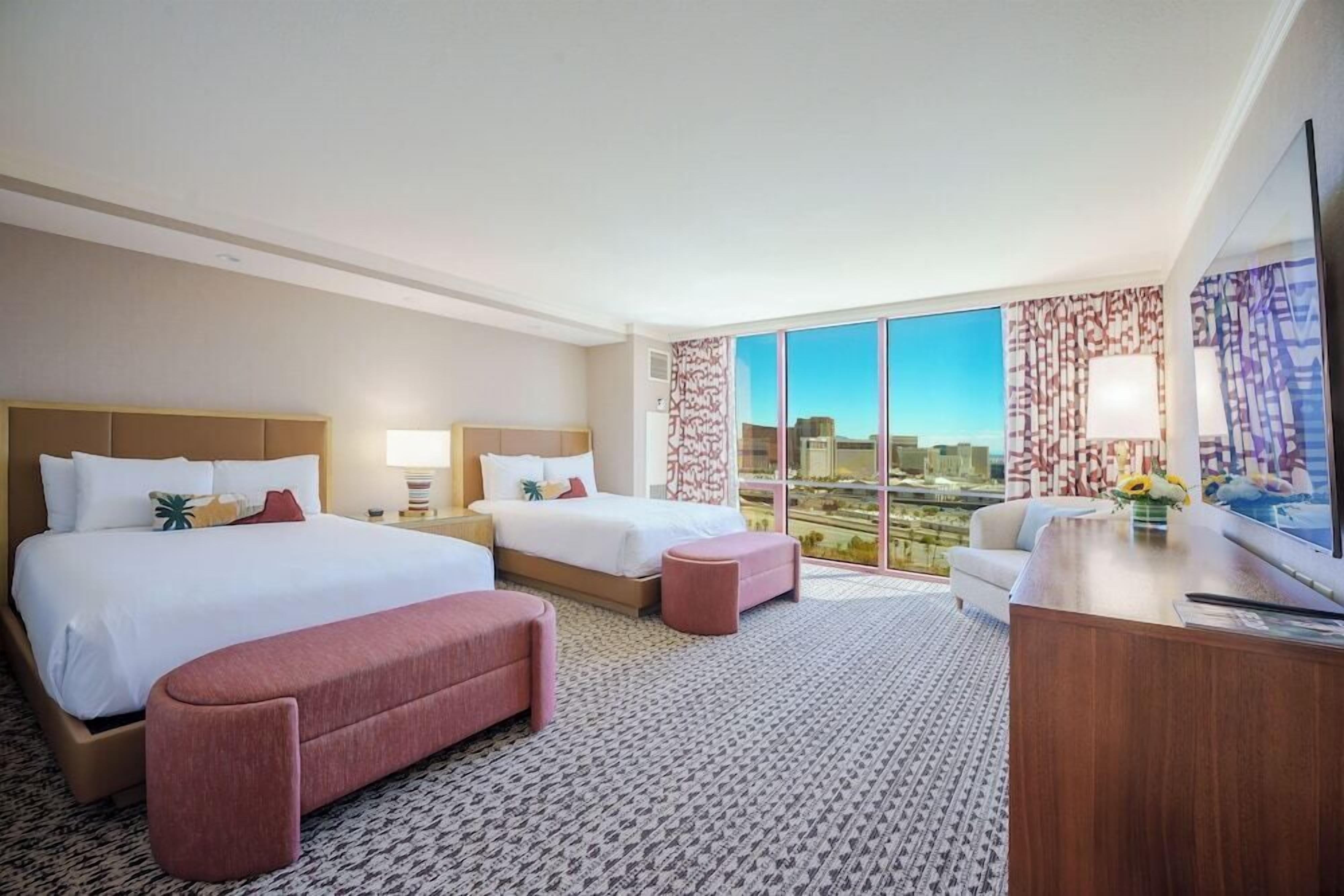 Ein Hotelzimmer mit zwei Betten, einem Schreibtisch, einem großen Fenster mit Blick auf die Stadt und einem gemusterten Teppich.