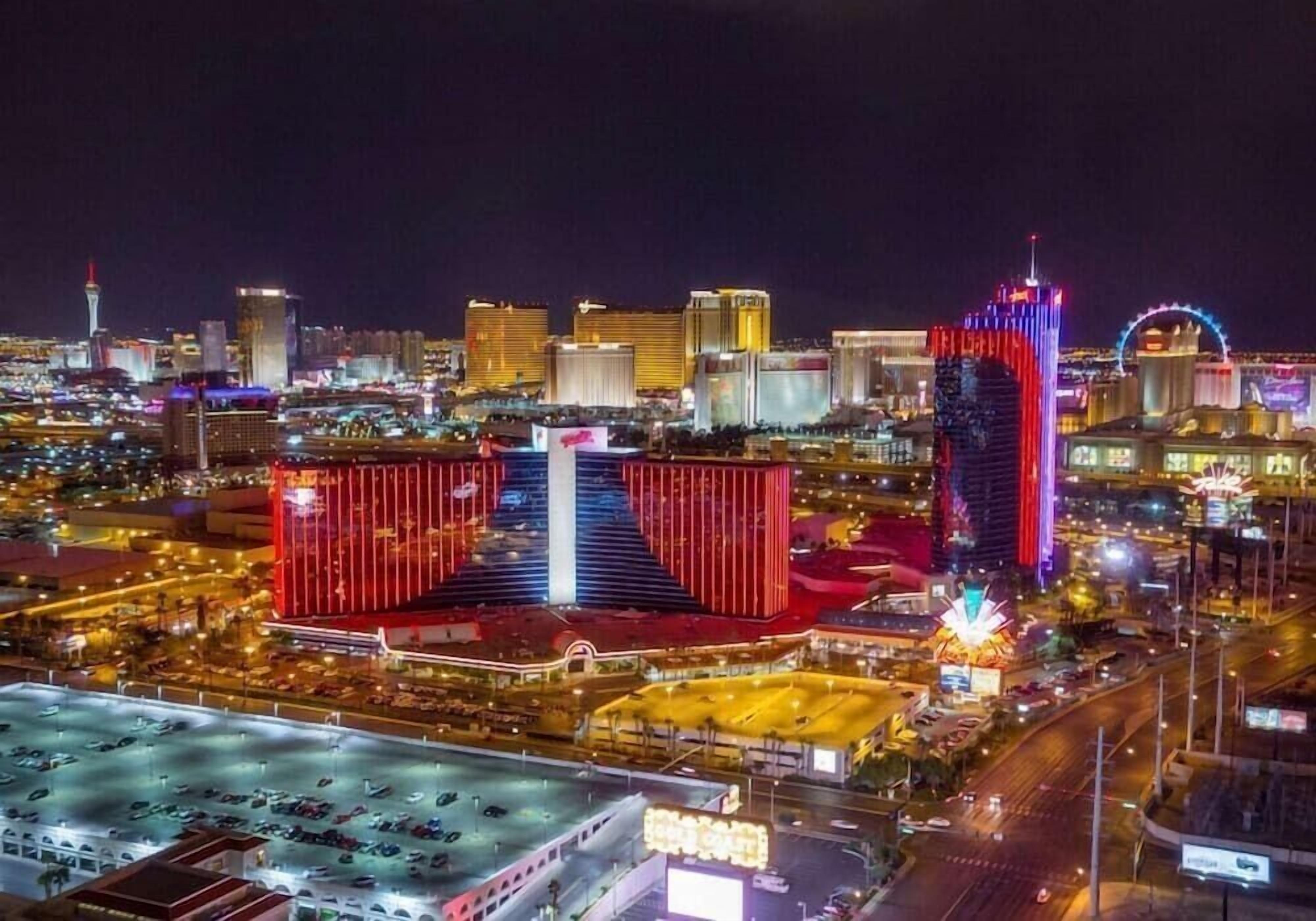 Une vue nocturne d’une ville, avec des bâtiments illuminés, un casino imposant aux éclairages rouge et bleu, et une grande roue visible au loin.