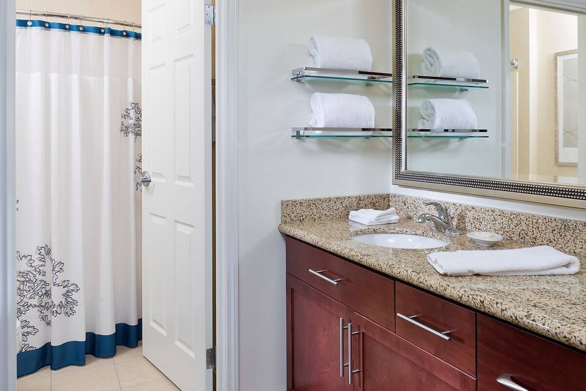 A bathroom with a shower curtain, a glass shelf with towels, a sink, and a mirror.