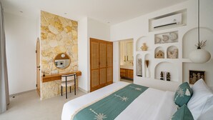A modern bedroom with a stone accent wall, a wooden desk, a large bed with green and white bedding, and a wall-mounted air conditioning unit. - Villa Maiora Tres Bingin (Pecatu)