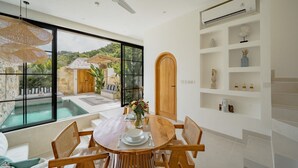 A dining area with a large window overlooking a pool and a view of a house with a thatched roof. - Villa Maiora Tres Bingin (Pecatu)