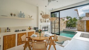 A modern kitchen with wooden cabinets, a dining area with a round table and chairs, a large window overlooking a pool, and a ceiling fan. - Villa Maiora Tres Bingin (Pecatu)