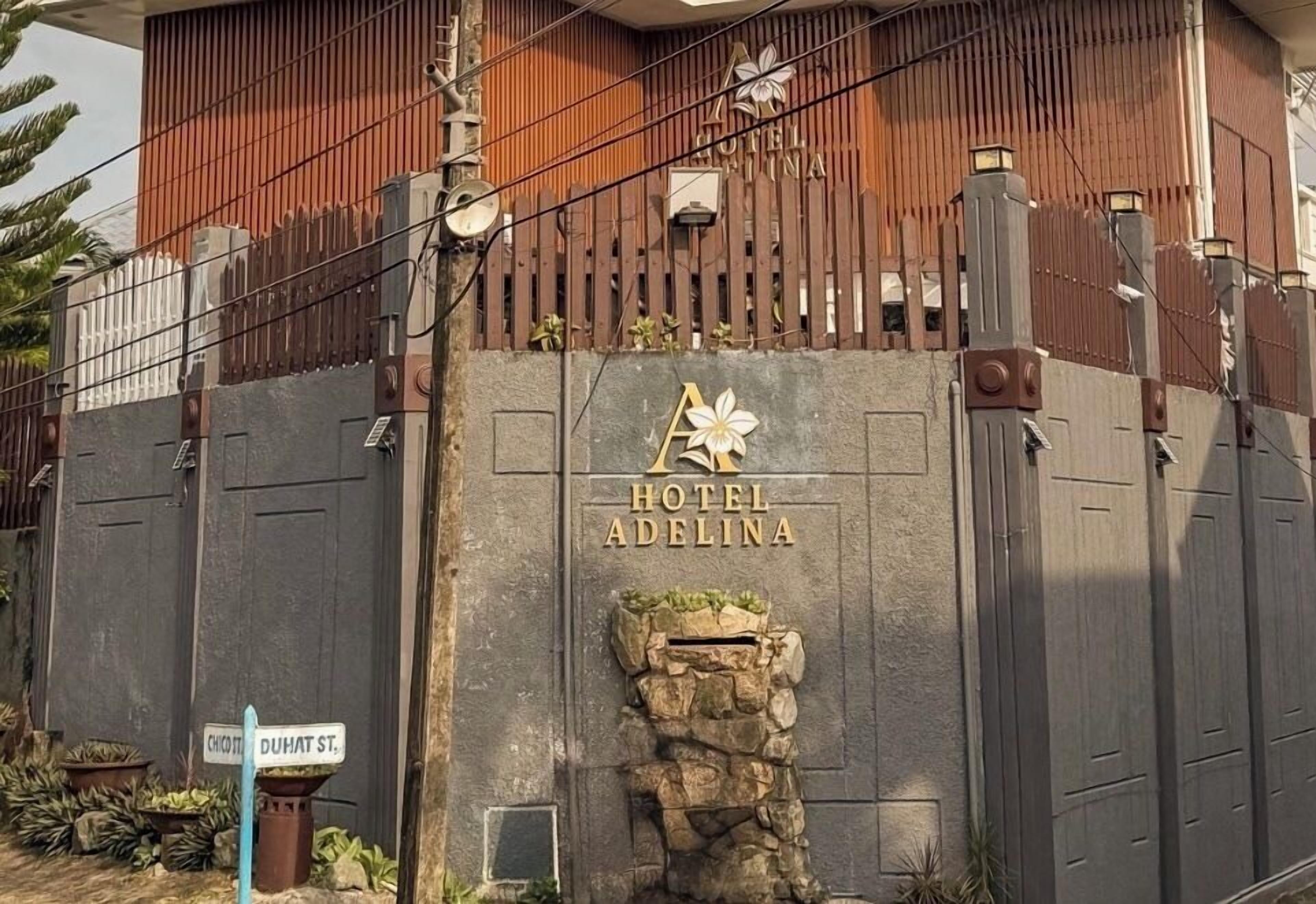 A hotel entrance with a stone facade, a wooden fence, and a sign indicating 'Hotel Adelina' and 'Choi Chung Dam St.'