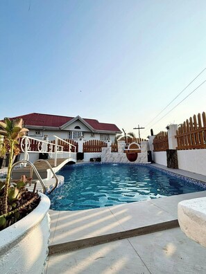 A swimming pool with a wooden fence, a white building with a red roof, and a palm tree. - Hotel Adelina Balanga City Bataan (Balanga)