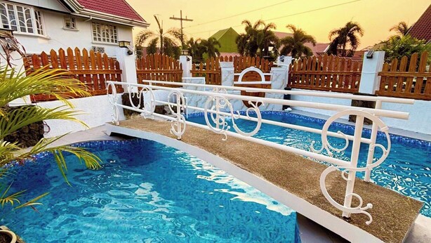 A swimming pool with a wooden fence, a white house with a red roof, and palm trees in the background.