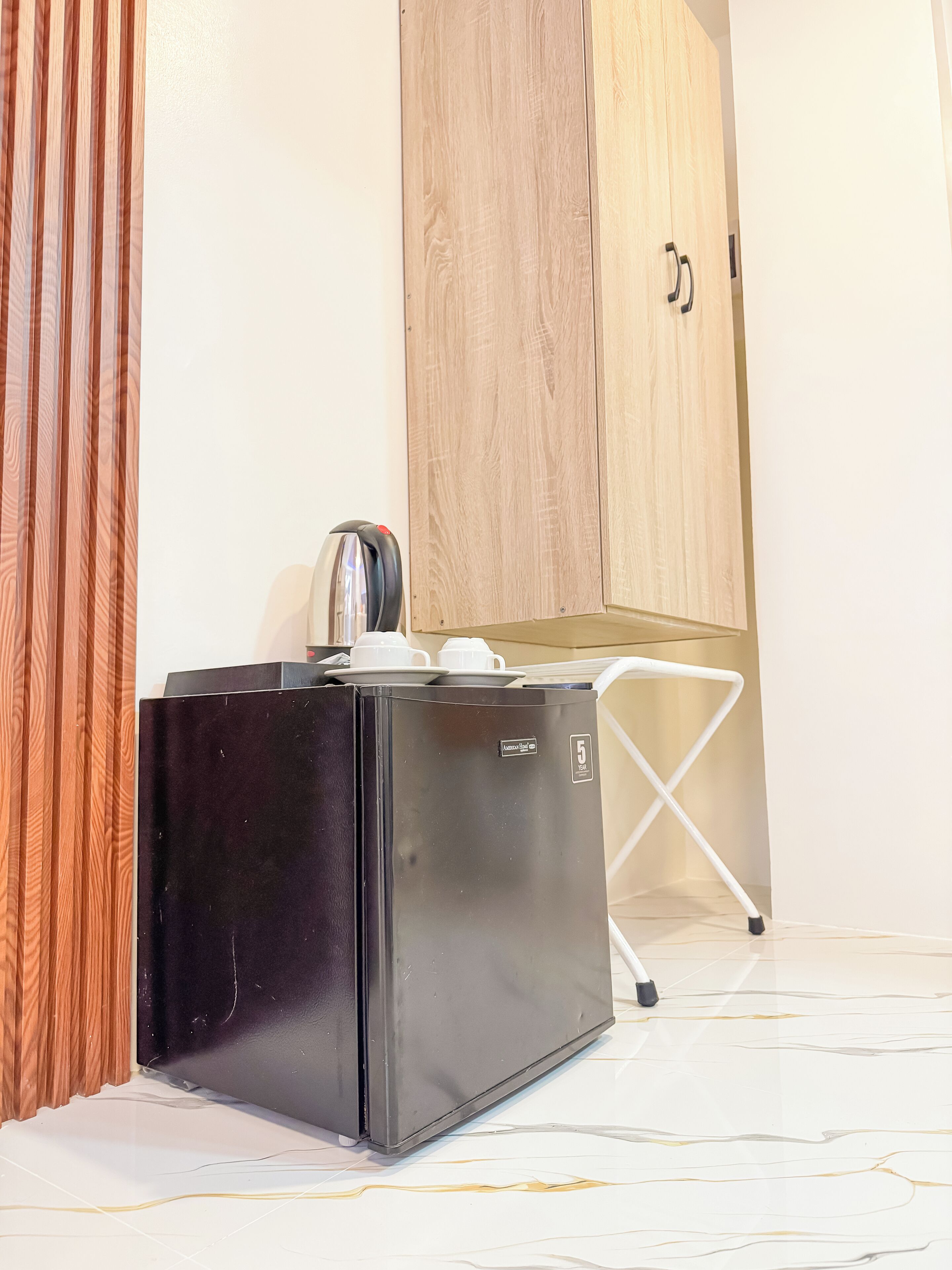 Un refrigerador negro compacto con una cafetera encima, una silla blanca plegable y un armario de madera al fondo.