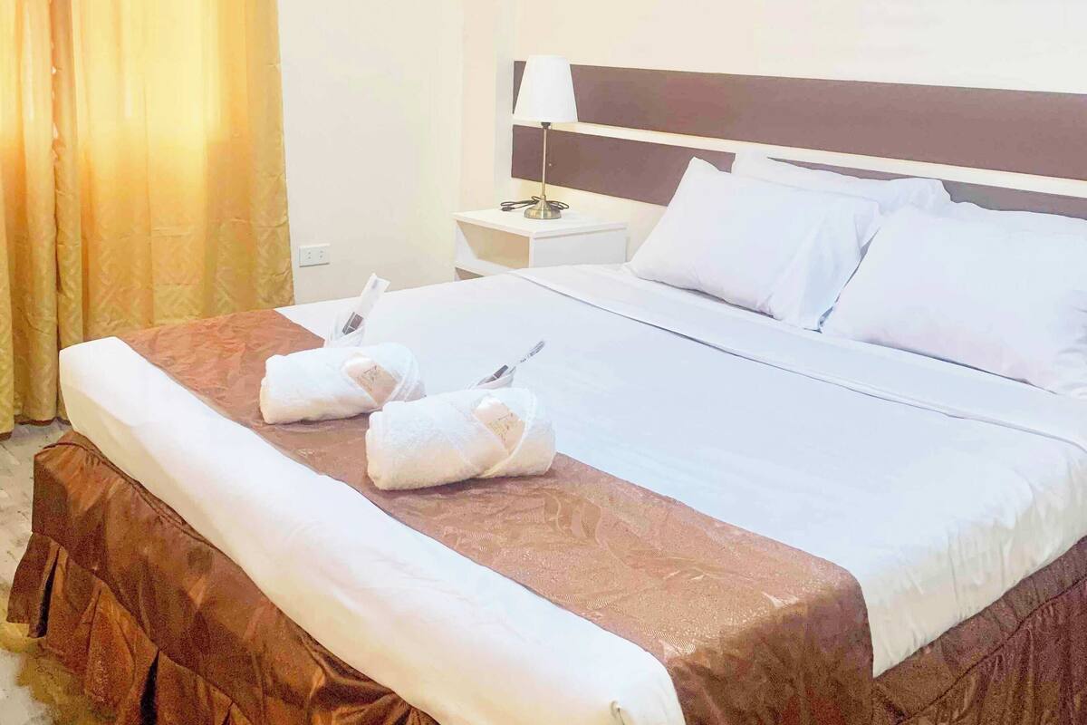 Ein Hotelzimmer mit einem ordentlich bezogenem Bett, einem Nachttisch mit Lampe und einem Fenster mit Vorhängen.