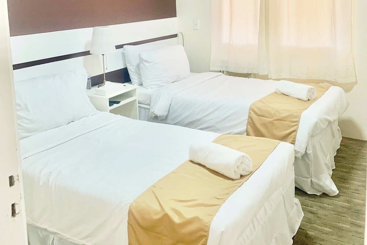 A hotel room with two beds, each with white linens and a beige bed runner. There is a nightstand with a lamp and a phone.