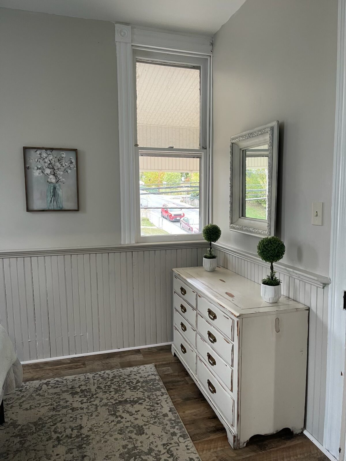 A room with a white dresser, a framed picture on the wall, and a window with a view of the street.