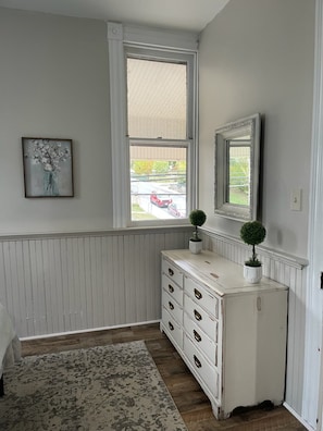 A room with a white dresser, a framed picture on the wall, and a window with a view of the street.