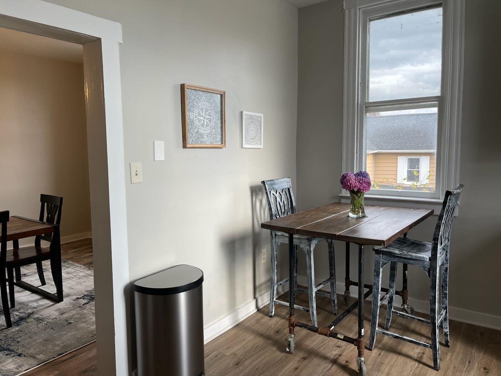 A dining area with a wooden table and chairs, a trash can, and two framed wall art pieces.