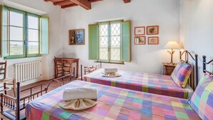 Una habitación con dos camas, una silla de madera, una mesa y un ventanal con contraventanas verdes.