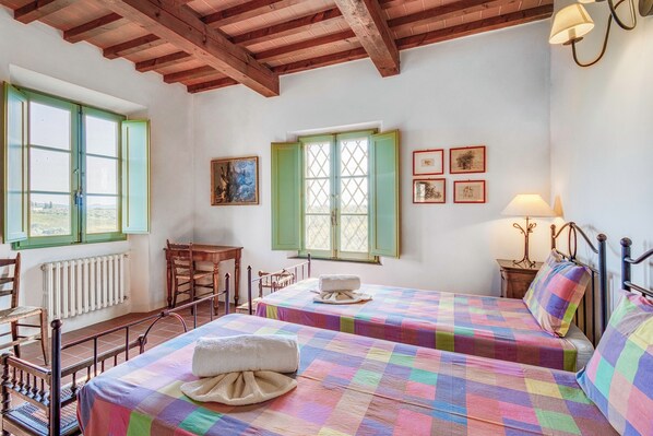 Quarto com duas camas, uma cadeira de madeira, uma mesa e uma janela com persianas verdes.