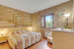 A bedroom with a stone wall, a bed with a striped bedspread, a bedside lamp, a window with a view, and a small table with a lamp and flowers. - Apt Ciclamino Shared Pool & Activities in Tuscany, Montaione, Italy (Montaione)