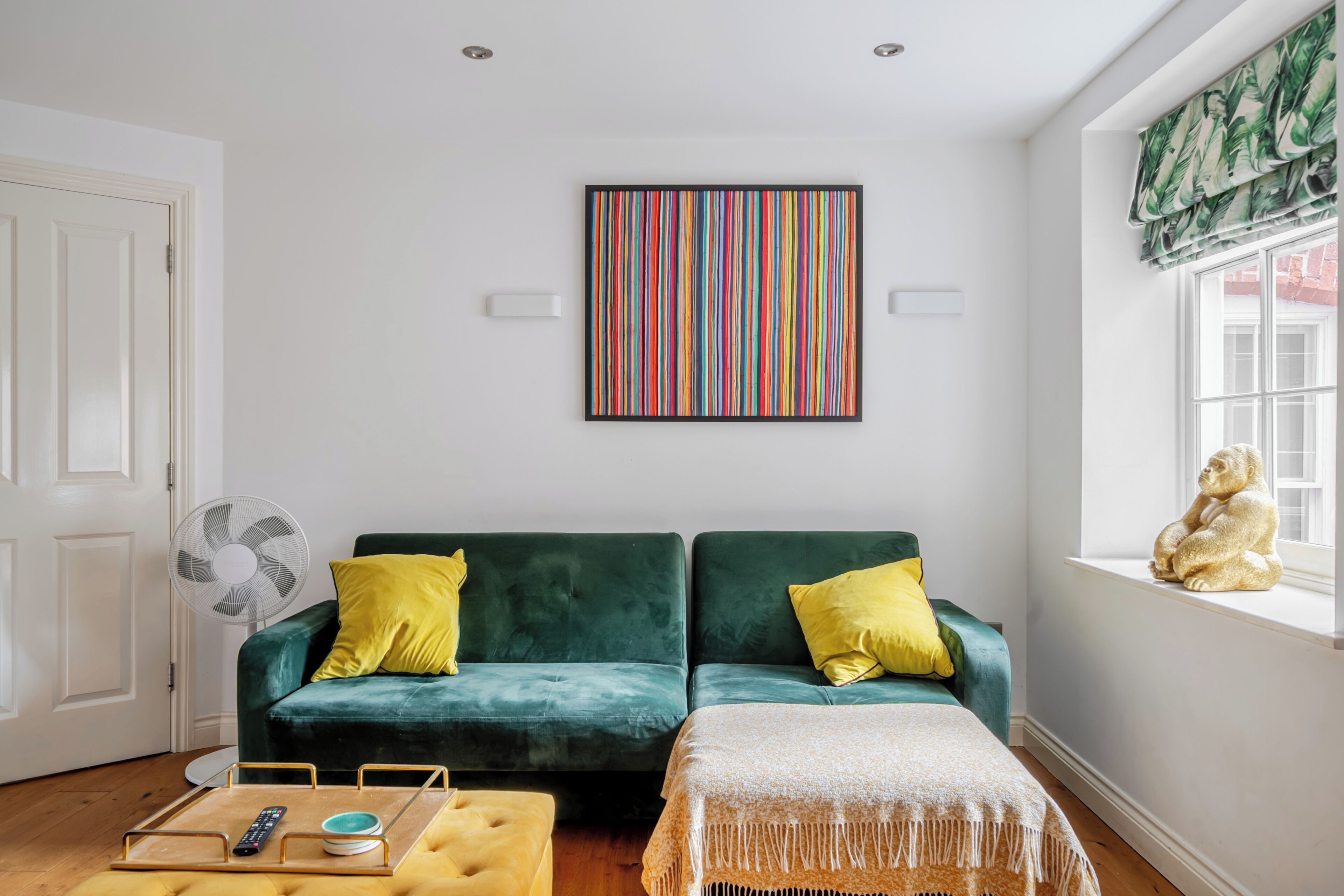 A room with a green sofa, yellow pillows, a framed artwork, a window with a view, and a small statue.