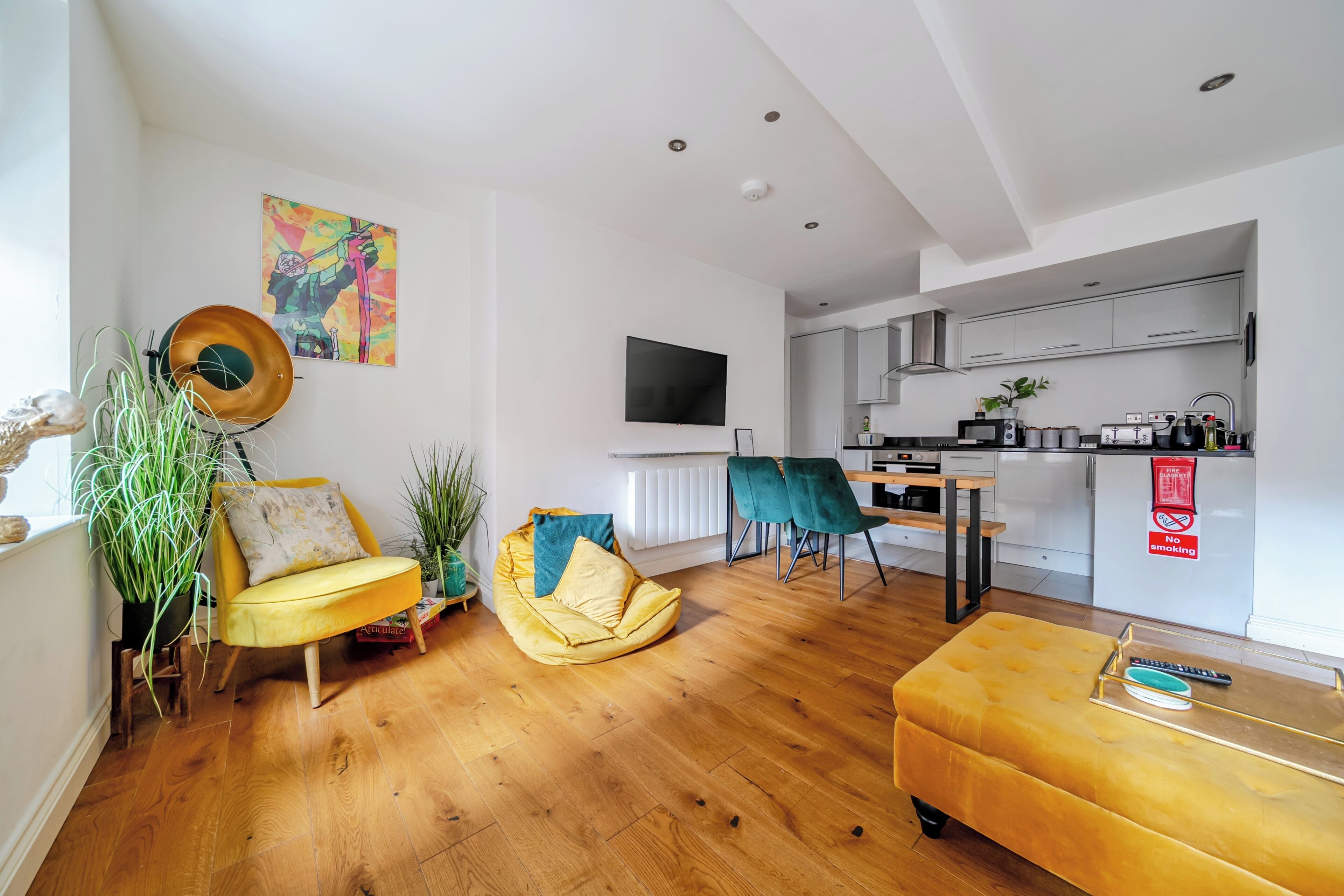 A modern living room with a yellow sofa, a wooden coffee table, and a green chair. A television is mounted on the wall, and a kitchen is visible in the background.