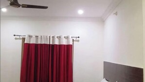 Una cama doble con sábanas blancas, un ventilador de techo y una ventana con cortinas rojas.