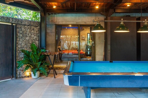 Une table de billard dans une pièce avec un mur en pierre, une grande plante en pot et, à travers une porte vitrée, on aperçoit un lit.