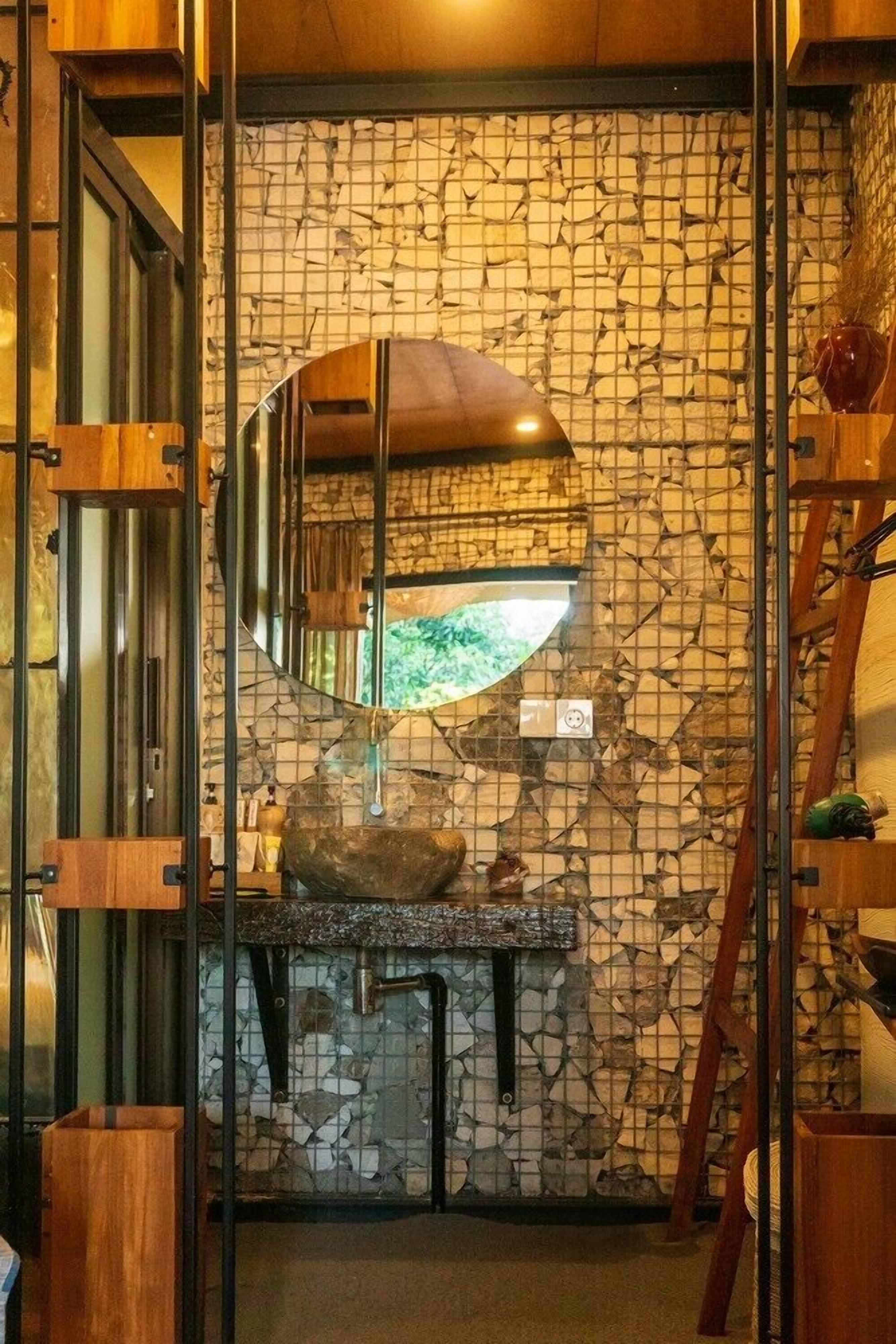 A bathroom with a stone mosaic wall, a wooden vanity, and a mirror reflecting a green outdoor view.