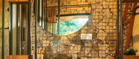 A bathroom with a stone mosaic wall, a wooden vanity, and a mirror reflecting a green outdoor view.