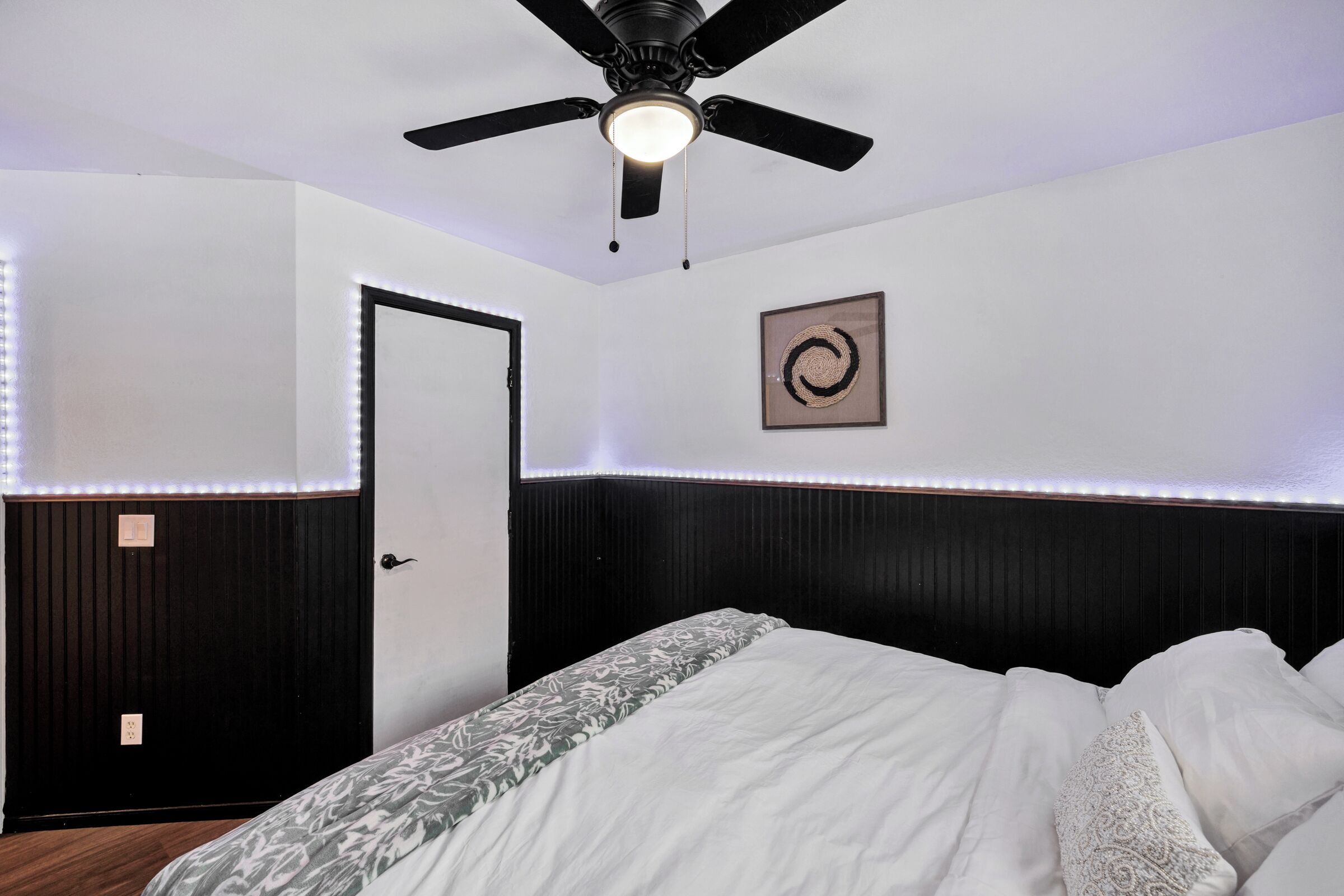 Une chambre à coucher avec un lit, un ventilateur de plafond, une porte et un tableau encadré au mur.