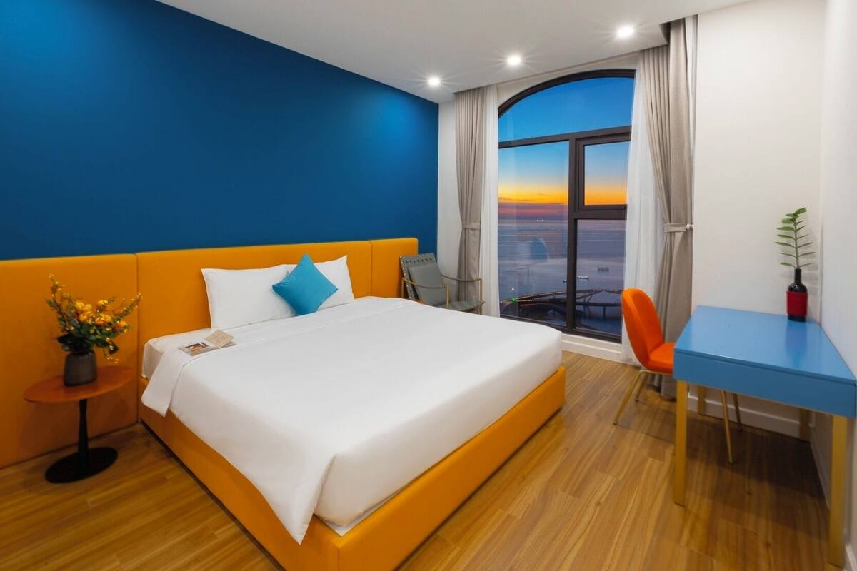Moderni hotellihuone, jossa on suuri sänky, työpöytä ja ikkunasta avautuva näkymä auringonlaskuun.