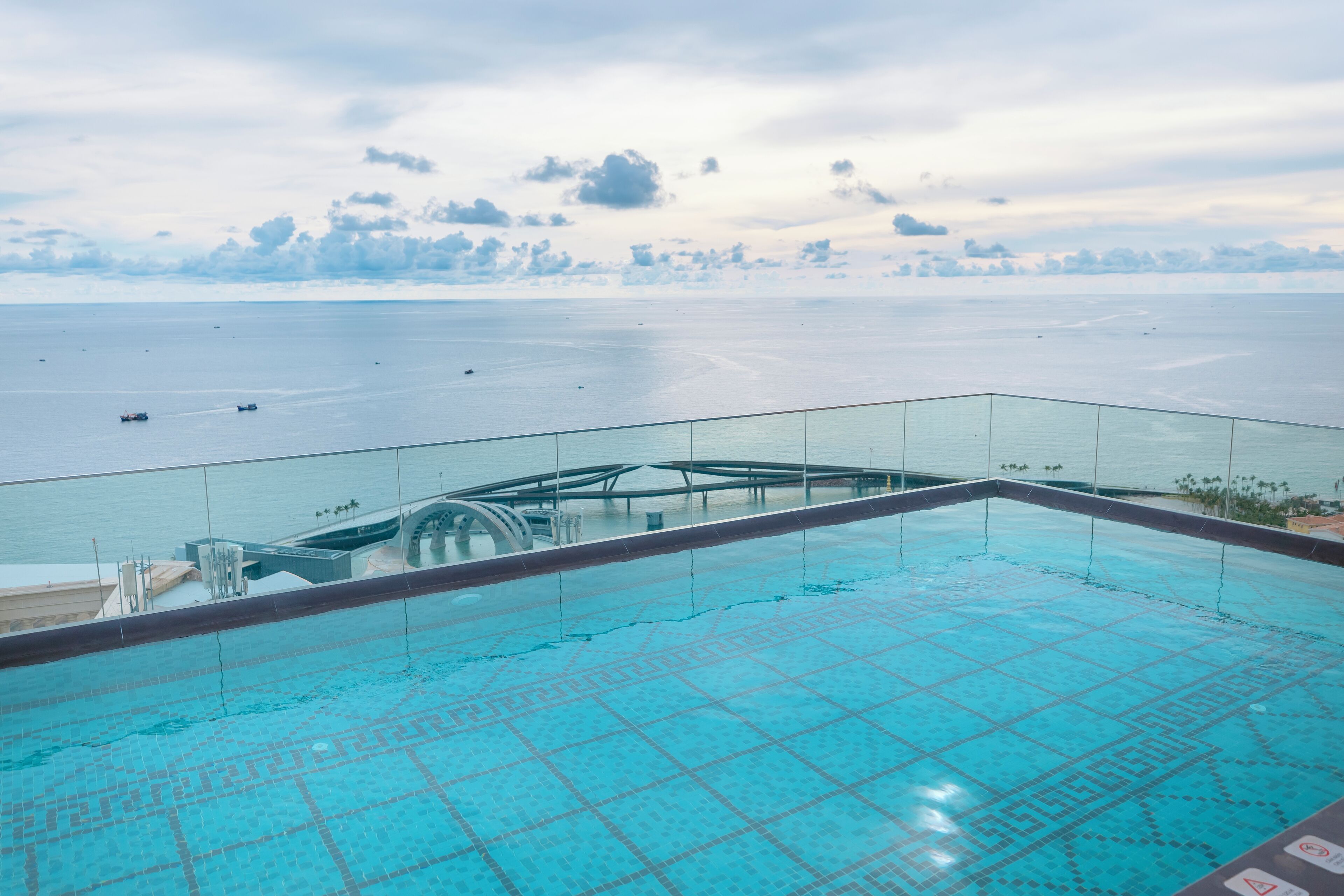 Ein Infinity-Pool auf dem Dach mit Glasgeländer, mit Blick auf ein ruhiges Meer und eine entfernte Küstenlinie.