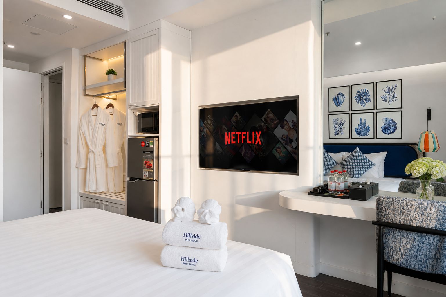 Habitación de hotel con cama, televisor mostrando 'NETFLIX', un minibar, una zona de estar con mesa y una pared decorada con obras de arte enmarcadas.