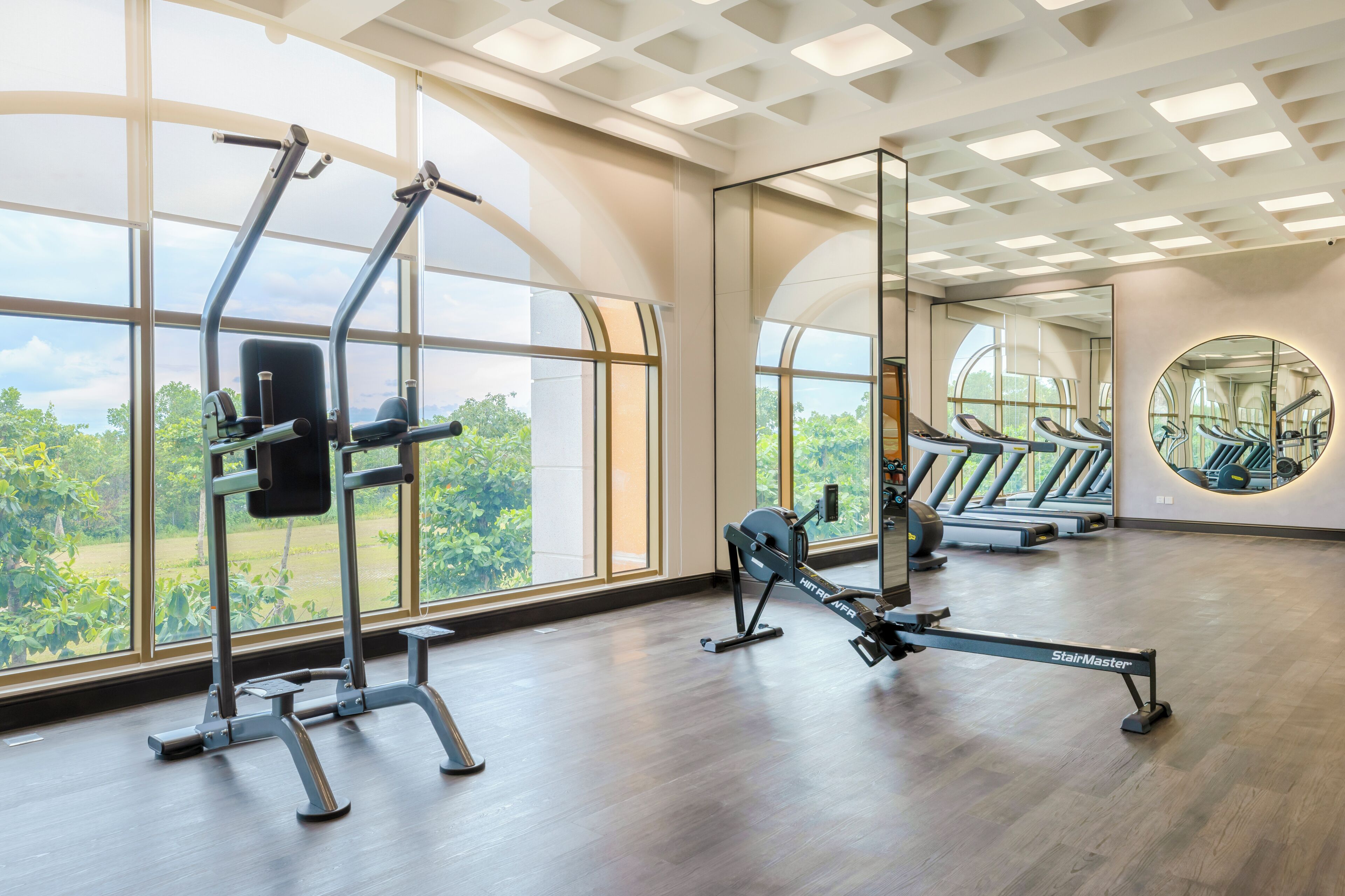 A gym with cardio machines, mirrored walls, and large windows offering a view of greenery.