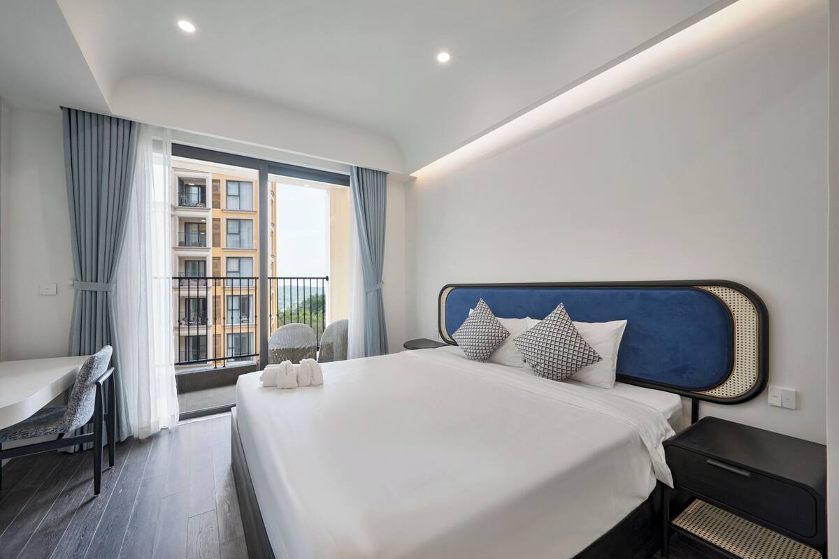 Hotellihuone, jossa on suuri sänky, työpöytä ja ikkunasta avautuva näkymä rakennukseen.