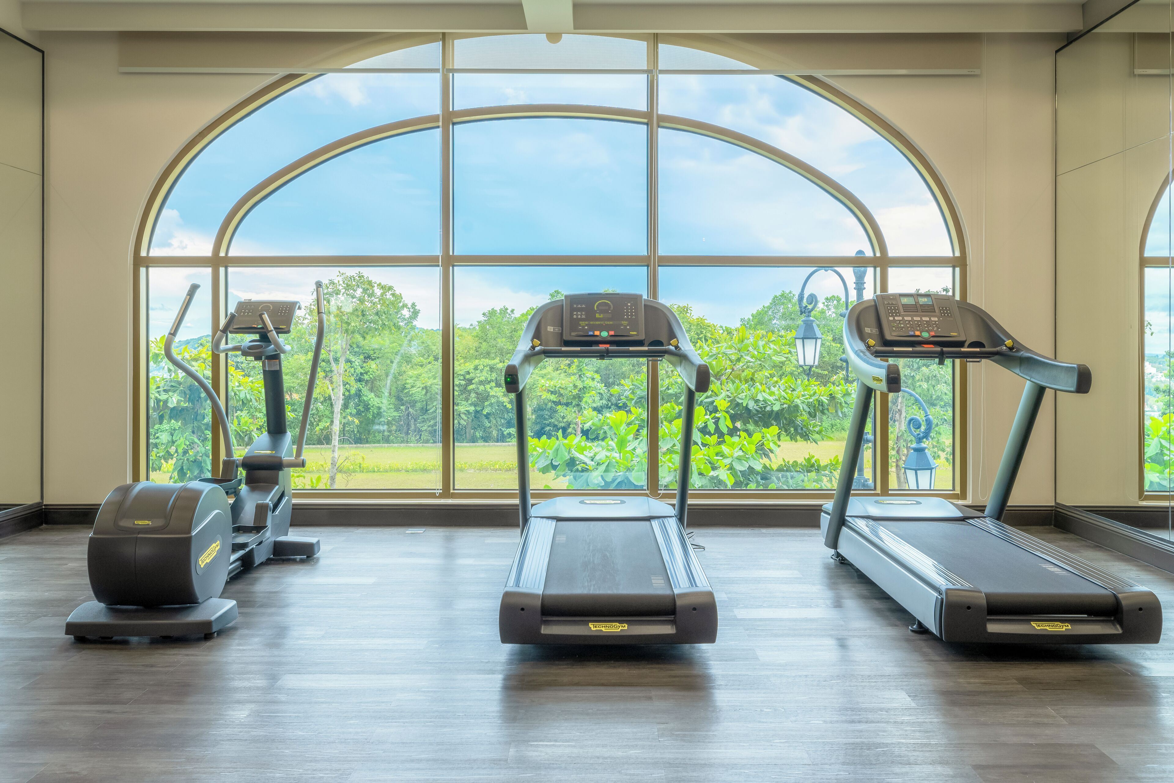 A gym with treadmills and elliptical machines, large windows with a view of greenery.