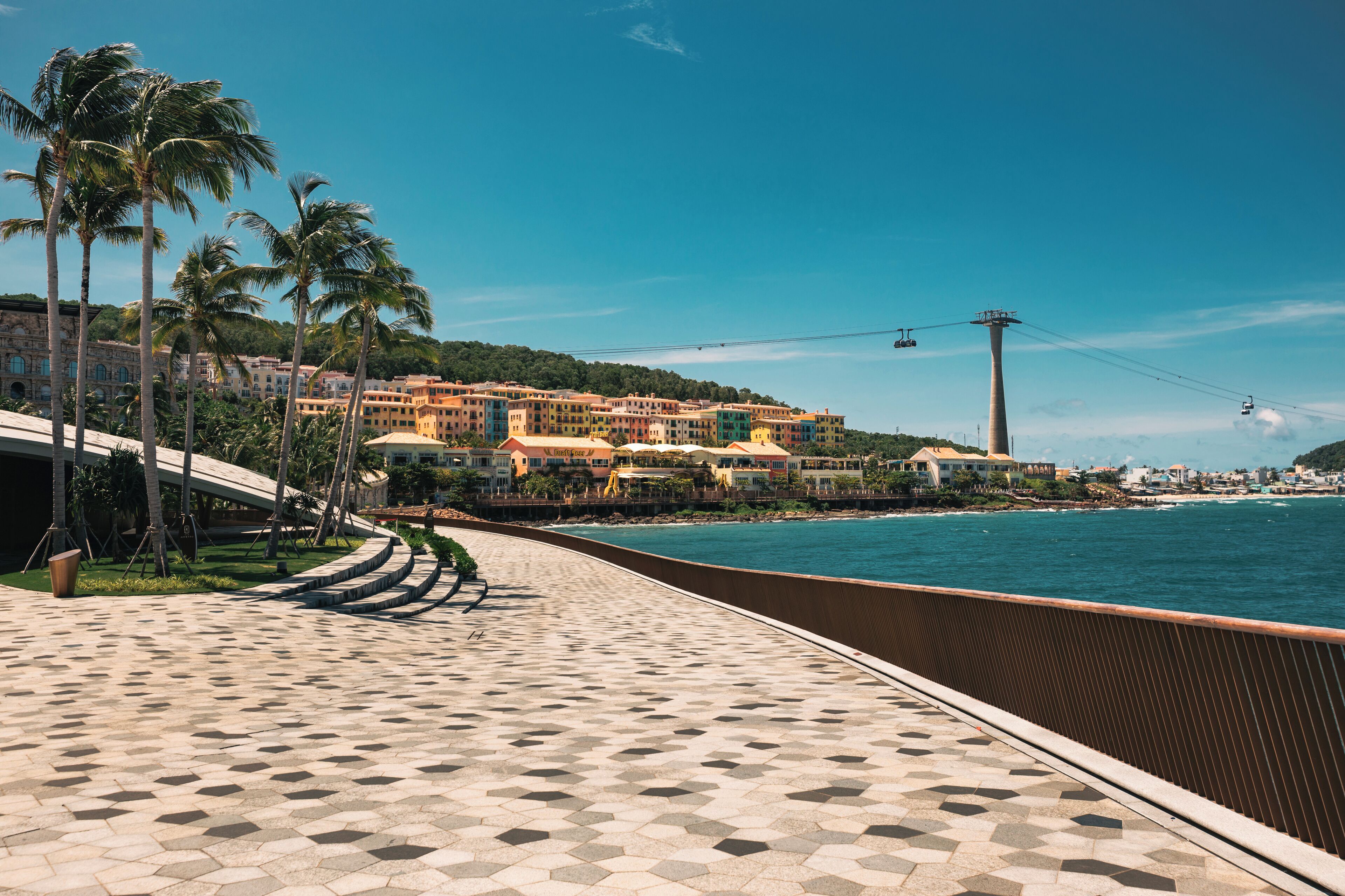 A coastal promenade with a tiled walkway, palm trees, and a row of colorful buildings with a view of the sea.