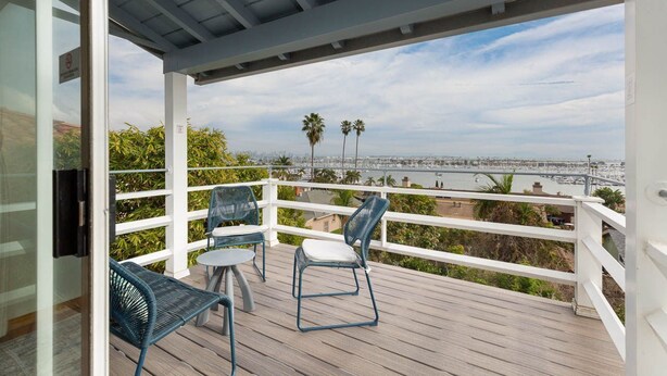 Een balkon met uitzicht op de oceaan en een brug, met een witte balustrade en blauwe stoelen.