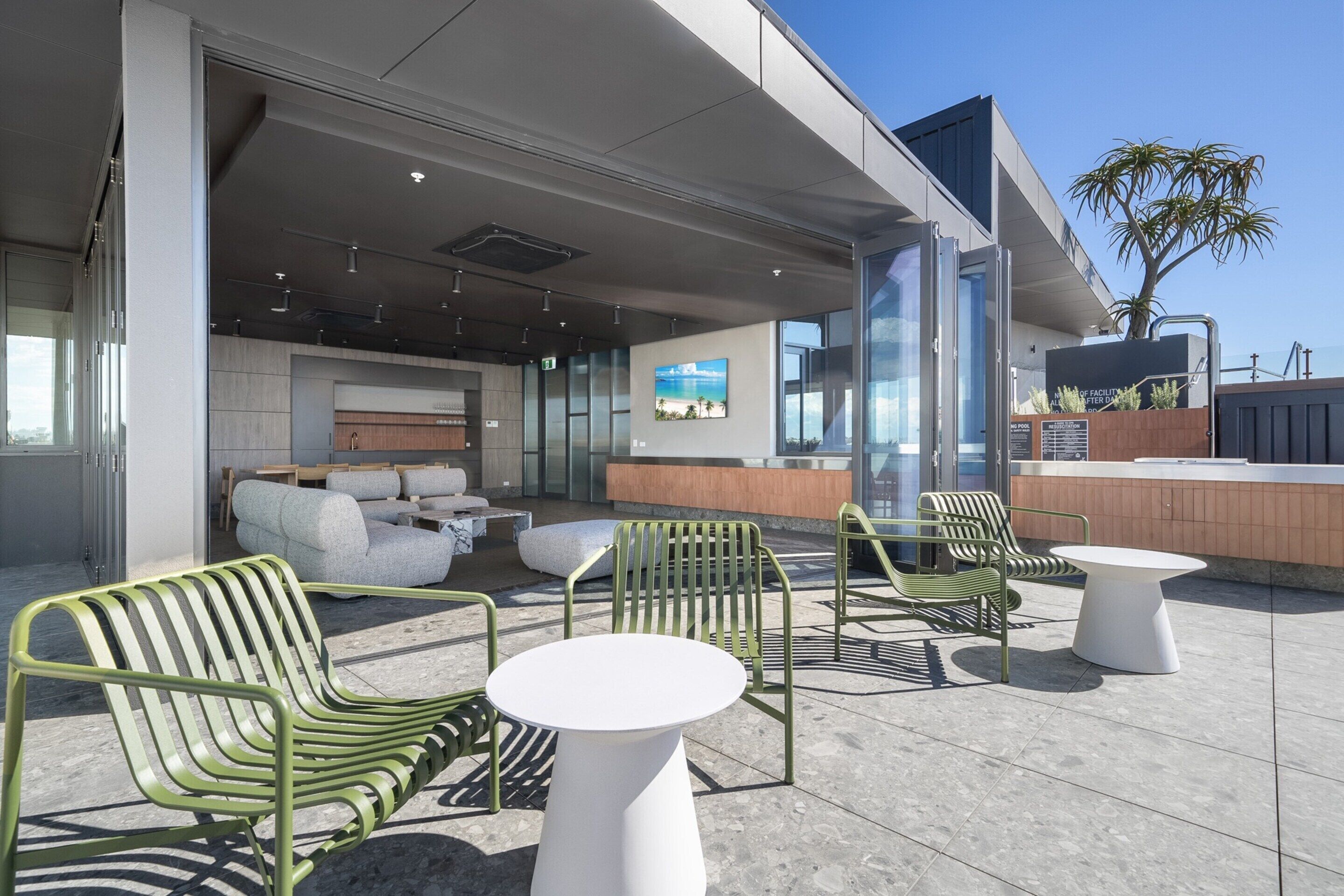An outdoor seating area with green chairs and white tables, a reception desk, and a large window with a view of the ocean.