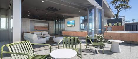 An outdoor seating area with green chairs and white tables, a reception desk, and a large window with a view of the ocean.